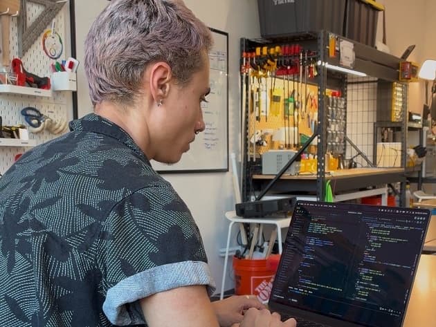 A maker coding on a laptop at a workbench in the Desk Job studio